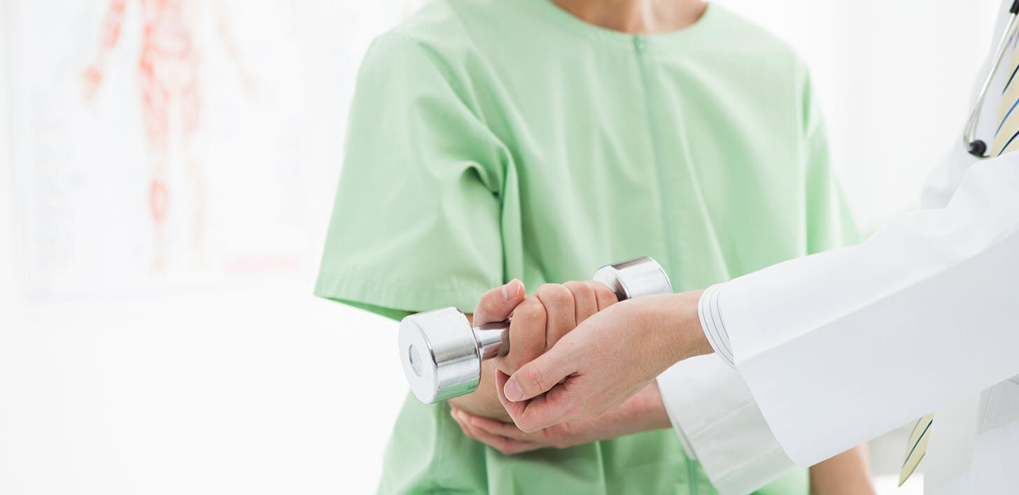 Pacjent w jasnozielonej bluzce trzymający hantel w ręku, ręce lekarza podtrzymujące hantel pacjenta.