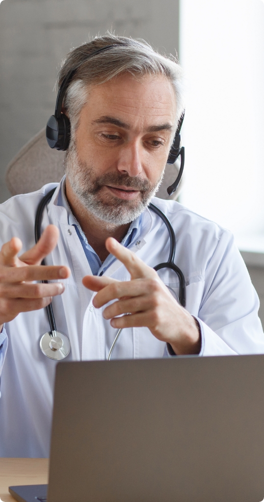 Lekarz w średnim wieku, z siwymi włosami i brodą, siedzacy przed laptopem i rozmawiający z kimś przez słuchawki.