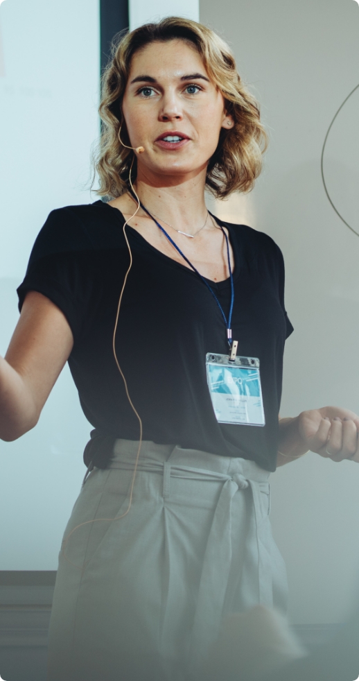 Młoda kobieta z blond srednimi włosami, prowadząca wykład podczas konferencji, ubrana w czarną bluzkę z krótkim rękawem i jasne spodnie.